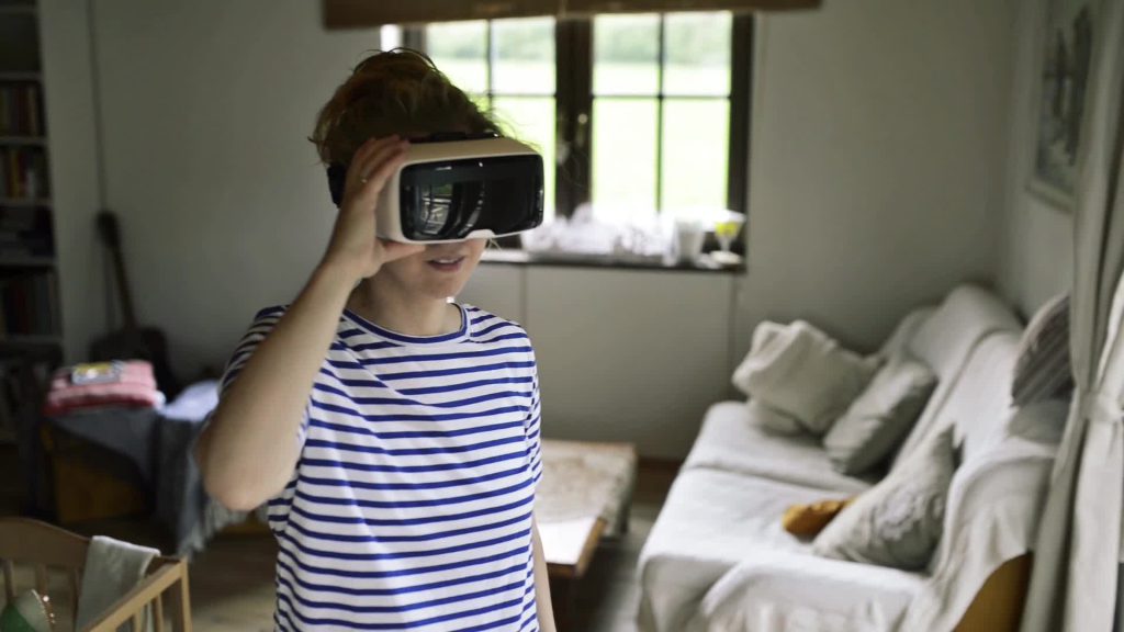 Young woman in blue striped t-shirt wearing virtual reality goggles standing in living room